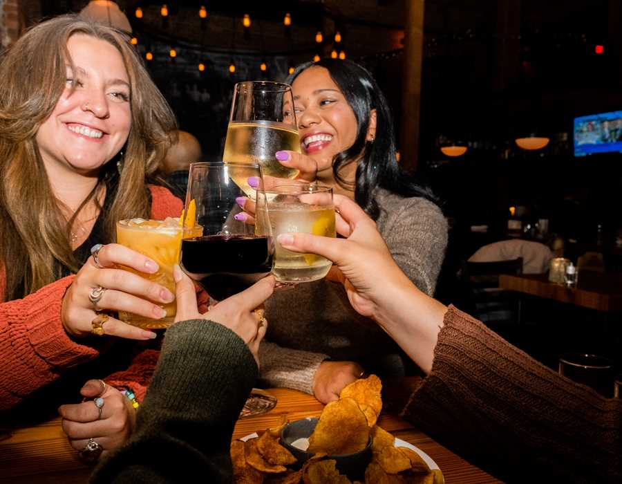 Four friends celebrate the end of the week doing a cheers with cocktails and wine over a table with chips and dip.